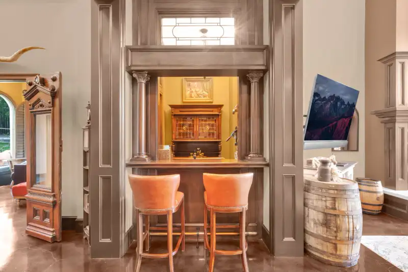Bar nook with two cognac leather barstools, paneled columns, and a barrel beside a wall-mounted television.