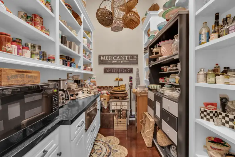 Walk-in pantry with white shelving, stocked jars and canned goods, woven baskets, and a wall sign reading Mercantile.