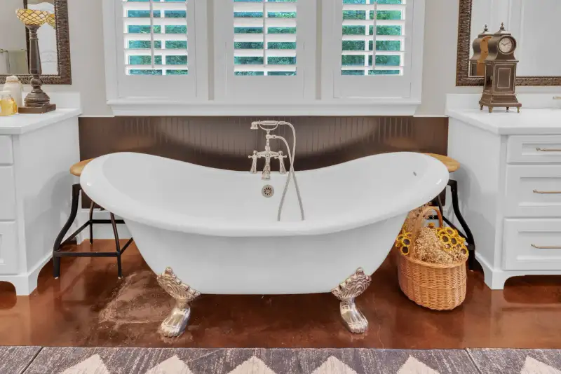 Clawfoot tub with silver feet beneath shuttered windows, beside white vanity cabinets and a wicker basket.