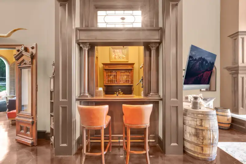 Wood-paneled bar nook with two tan leather stools, wood columns, a wine barrel, and a wall-mounted television.