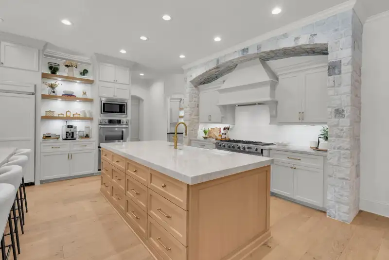 Kitchen with a light wood island, a stone arch over a white range hood, and white cabinetry.