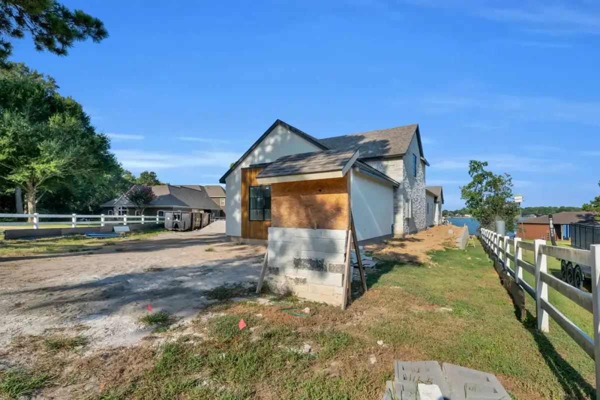 Side exterior of an unfinished stucco house with a block base and a white rail fence.