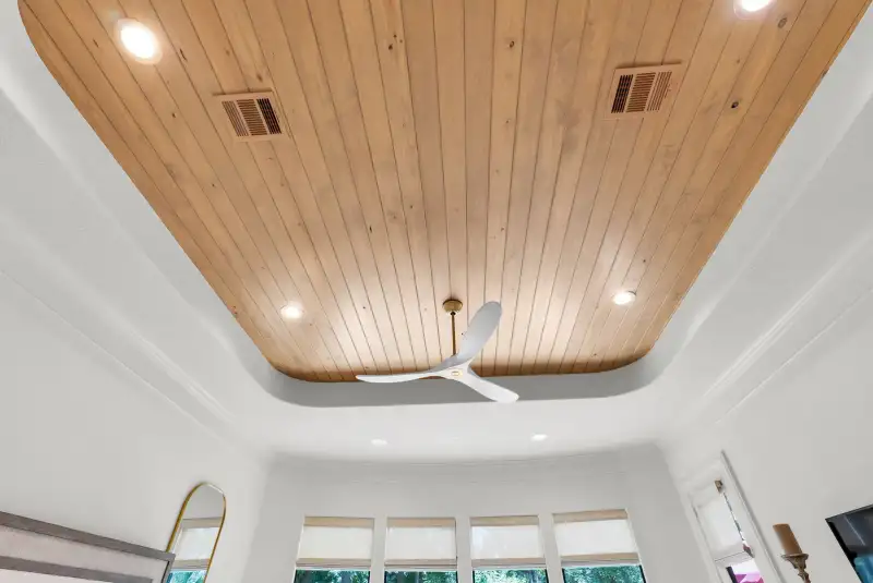 Looking up at a wood plank tray ceiling with recessed lights and a white ceiling fan.