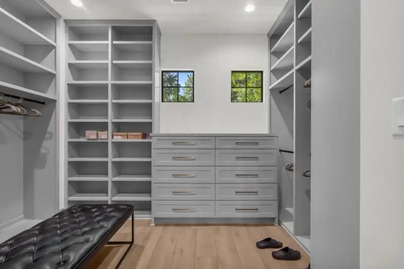 Walk-in closet with gray shelving, a tufted leather bench, a gray drawer unit, and two black-framed windows.