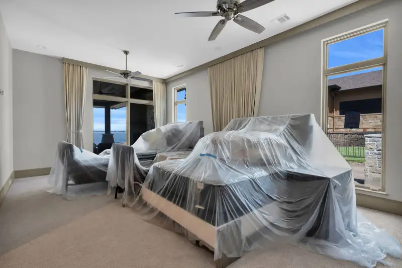 Bedroom with light walls, covered furniture, a ceiling fan, and two tall windows.