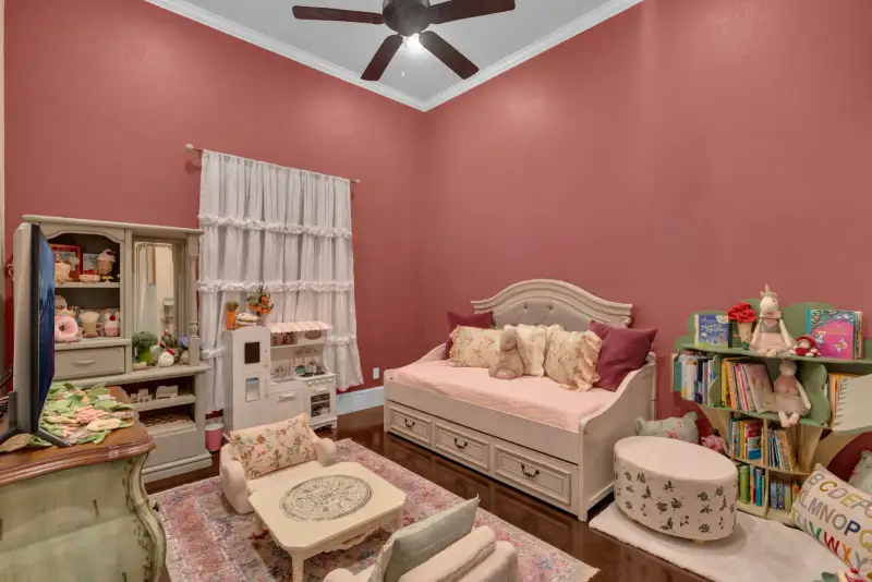 Child's room with rose-colored walls, a cream daybed, white ruffled curtains, and a green bookcase.