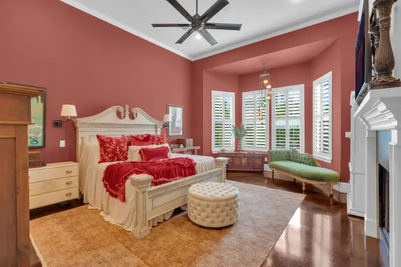 Bedroom with coral-red walls, a cream carved bed with red bedding, plantation shutters, and a green chaise.