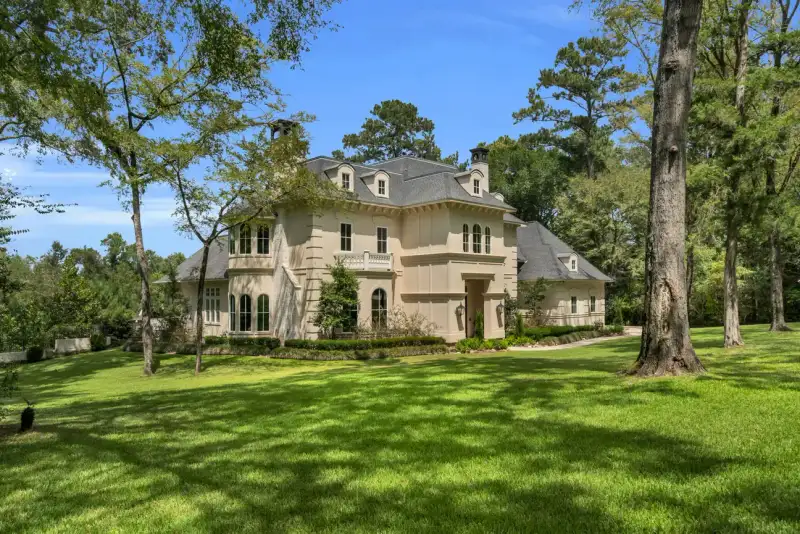 Cream stucco two-story home with a gray slate roof, surrounded by mature trees and a manicured front lawn.