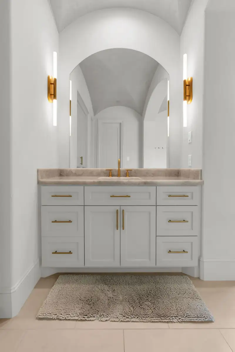 Single vanity with a marble-look top, an arched mirror, and brass wall sconces over a bath mat.
