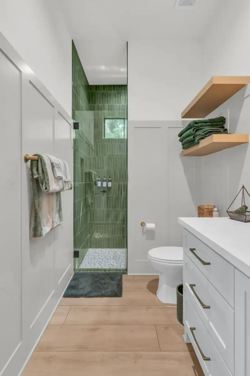 Bathroom with a green tiled shower, floating wood shelves, and a gray vanity beside a toilet.