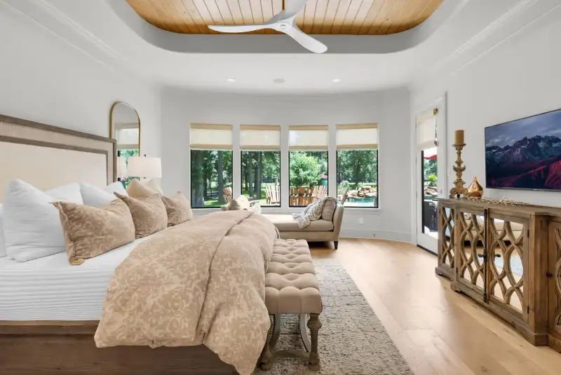 Bedroom with a bay of windows, a mirrored dresser, a wall-mounted television, and a tufted bench.