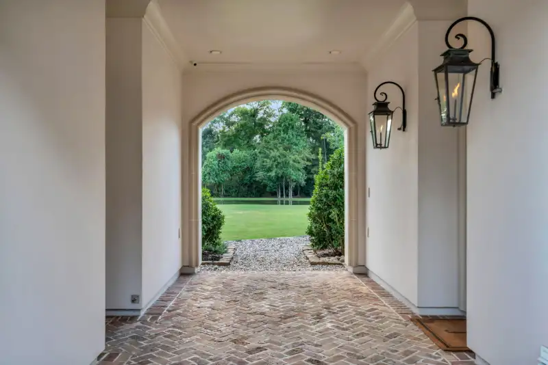 Arched brick passage with wall lanterns, looking through an opening to a lawn and trees.