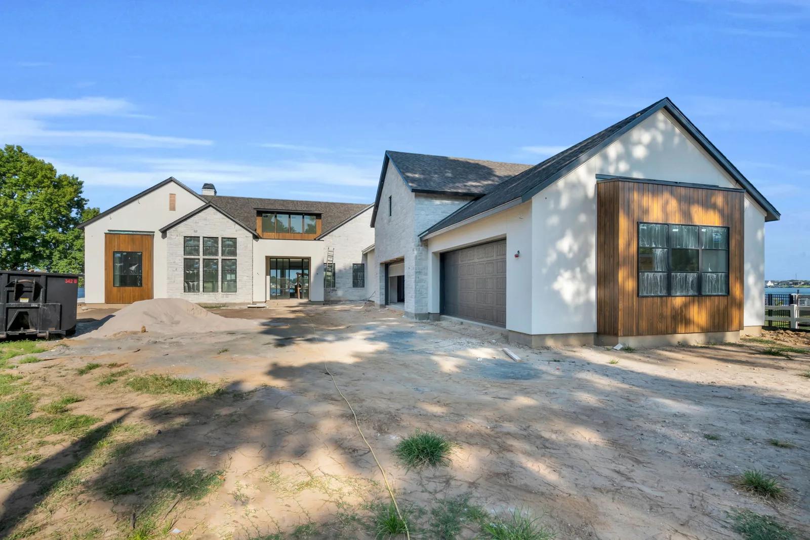 Front exterior of a stucco and stone house with wood accents and garage doors on a dirt drive.