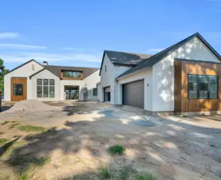 Front exterior of a stucco and stone house with wood accents and garage doors on a dirt drive.