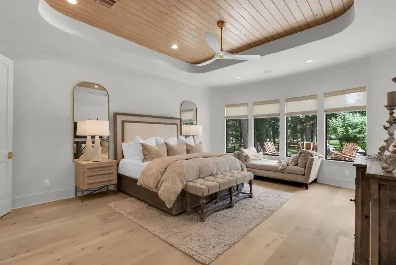 Primary bedroom with a wood plank tray ceiling, a ceiling fan, a bed, and an upholstered chaise by windows.