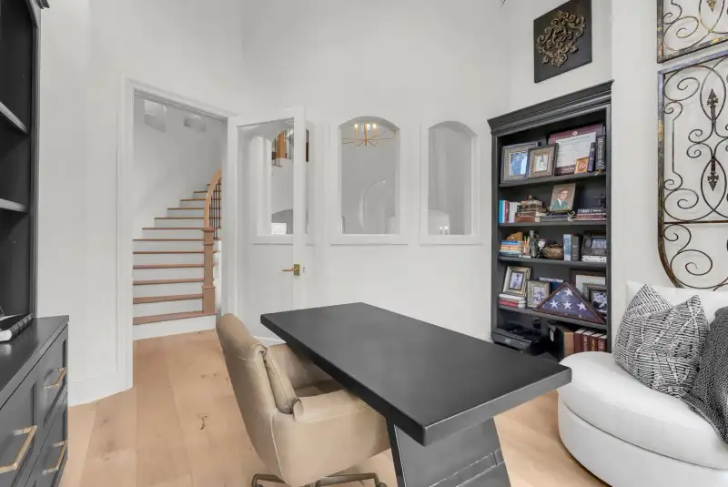 Study with a dark bookcase, framed photographs, and a dark desk near a staircase.