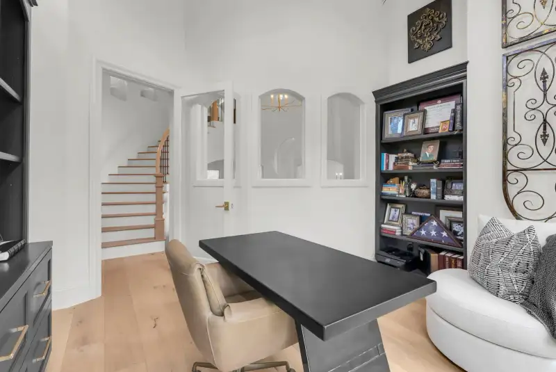 Study with a dark bookcase, framed photographs, and a dark desk near a staircase.