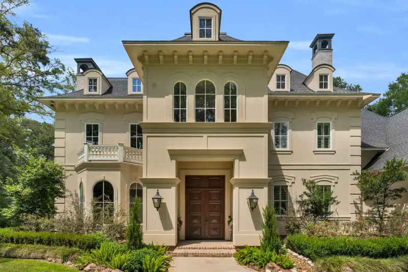 Front facade of a cream stucco home with a carved wood double door, brick entry steps, and clipped boxwood hedges.