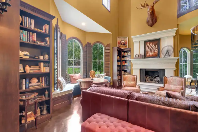 Two-story living room with yellow walls, bookcases, a white fireplace mantel, leather sofa, and arched windows.