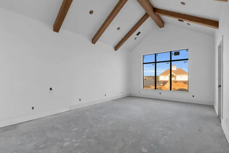 Vaulted room with exposed wood beams, a black-framed window, and bare concrete floors.