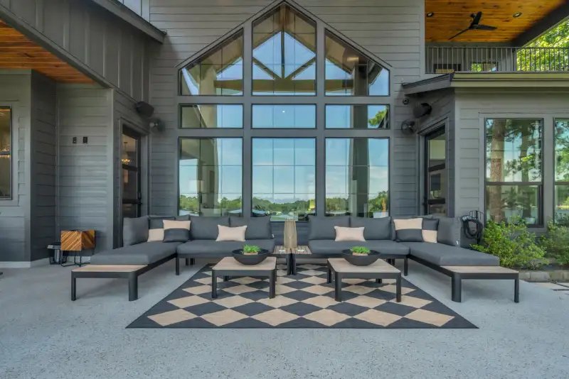 Covered outdoor living space with gray sectional seating and a checkered rug beneath a tall gabled window.