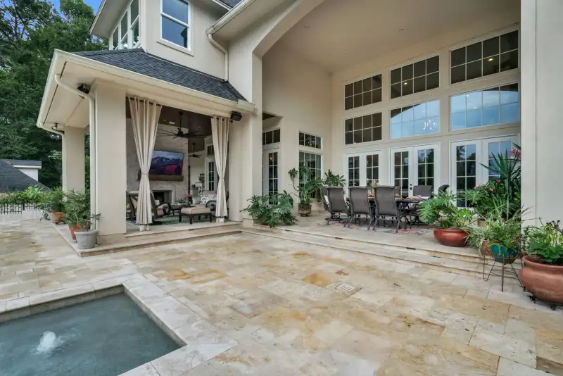 Covered patio with a dining table, tall divided-light windows, potted plants, and a travertine stone floor.