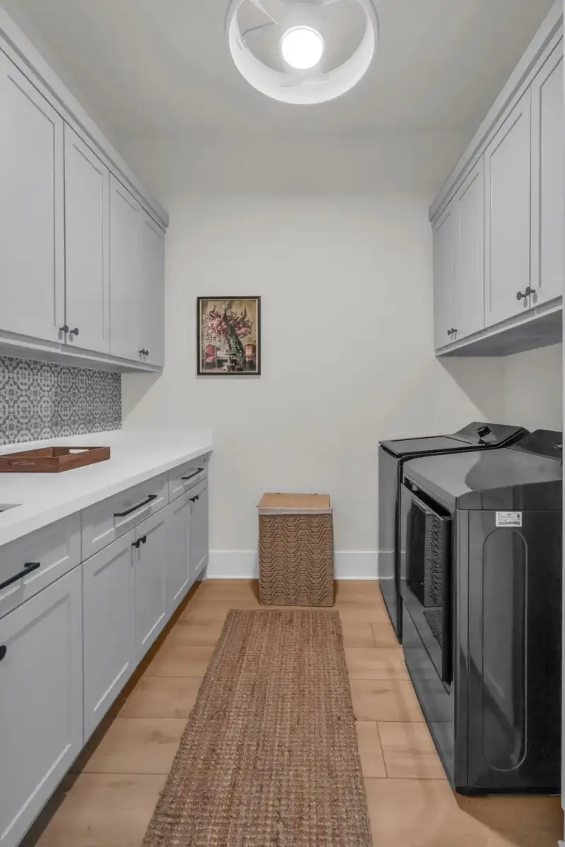 Laundry room with gray cabinets, a patterned tile backsplash, a black washer and dryer, and a woven runner rug.