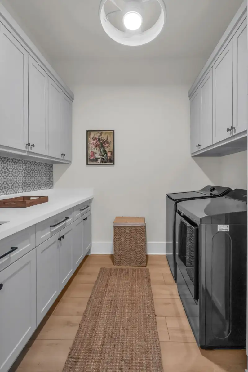 Laundry room with gray cabinets, a patterned tile backsplash, a black washer and dryer, and a woven runner rug.