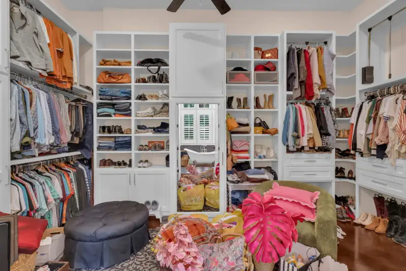Walk-in closet with white built-in shelving, hanging clothes, a tufted ottoman, and a mirrored closet door.