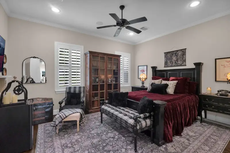 Bedroom with a ceiling fan, a dark wood armoire, a red velvet bed, and plantation shutters.