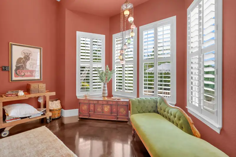 Sitting room with coral walls, white plantation shutters, a green tufted chaise, and a hanging glass pendant lamp.