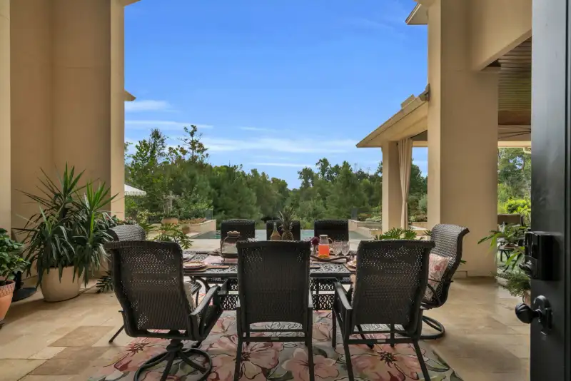 Covered outdoor dining area with a wicker chair set, a patterned rug, travertine floor, and a view of the pool.