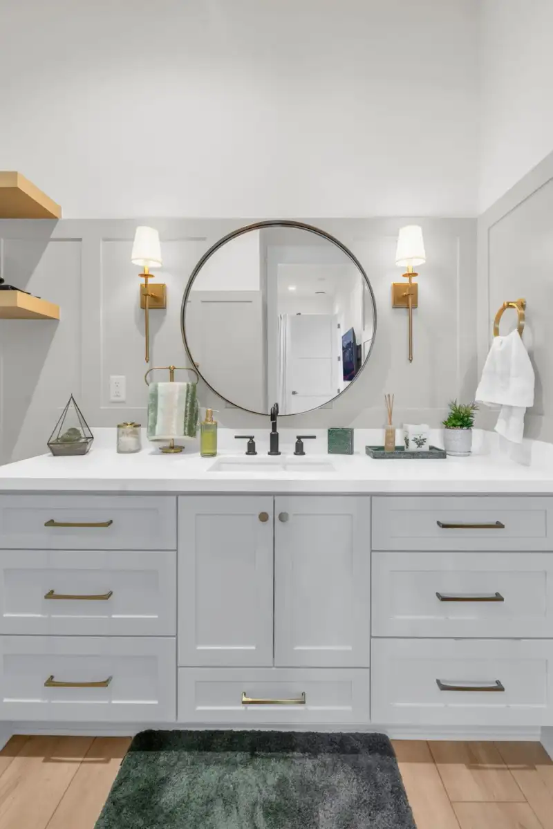 Bathroom vanity with gray cabinets, a round mirror, and brass wall sconces.