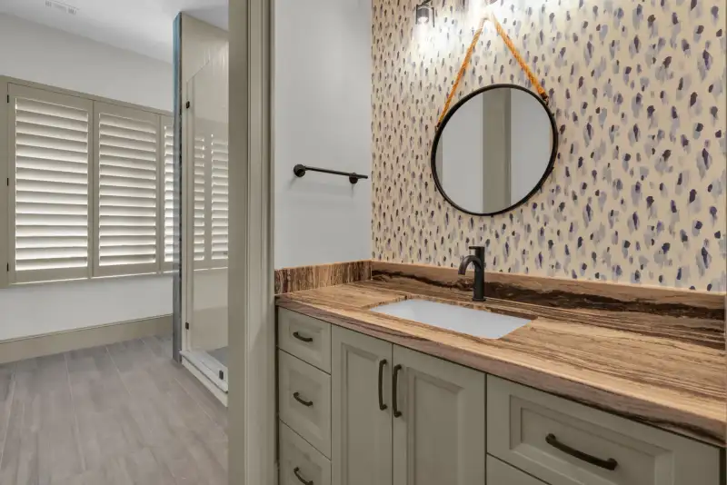 Powder room vanity with a round mirror, patterned wallpaper, and a veined brown countertop.