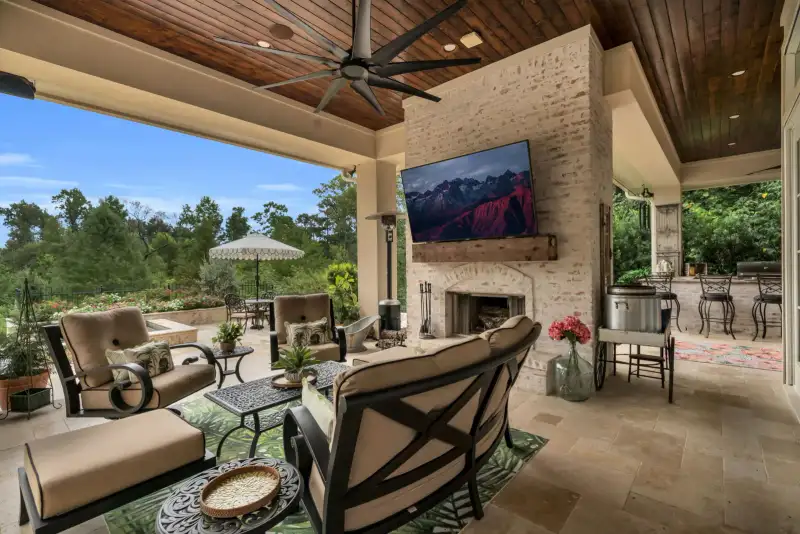 Covered patio with a brick fireplace, a wall-mounted television, cushioned seating, and a wood-planked ceiling.
