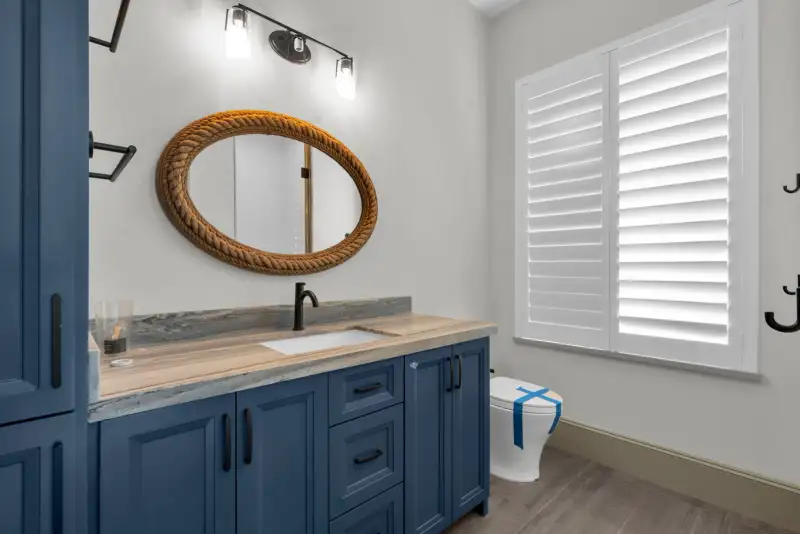 Bathroom with a blue vanity, a rope-framed oval mirror, and white plantation shutters.