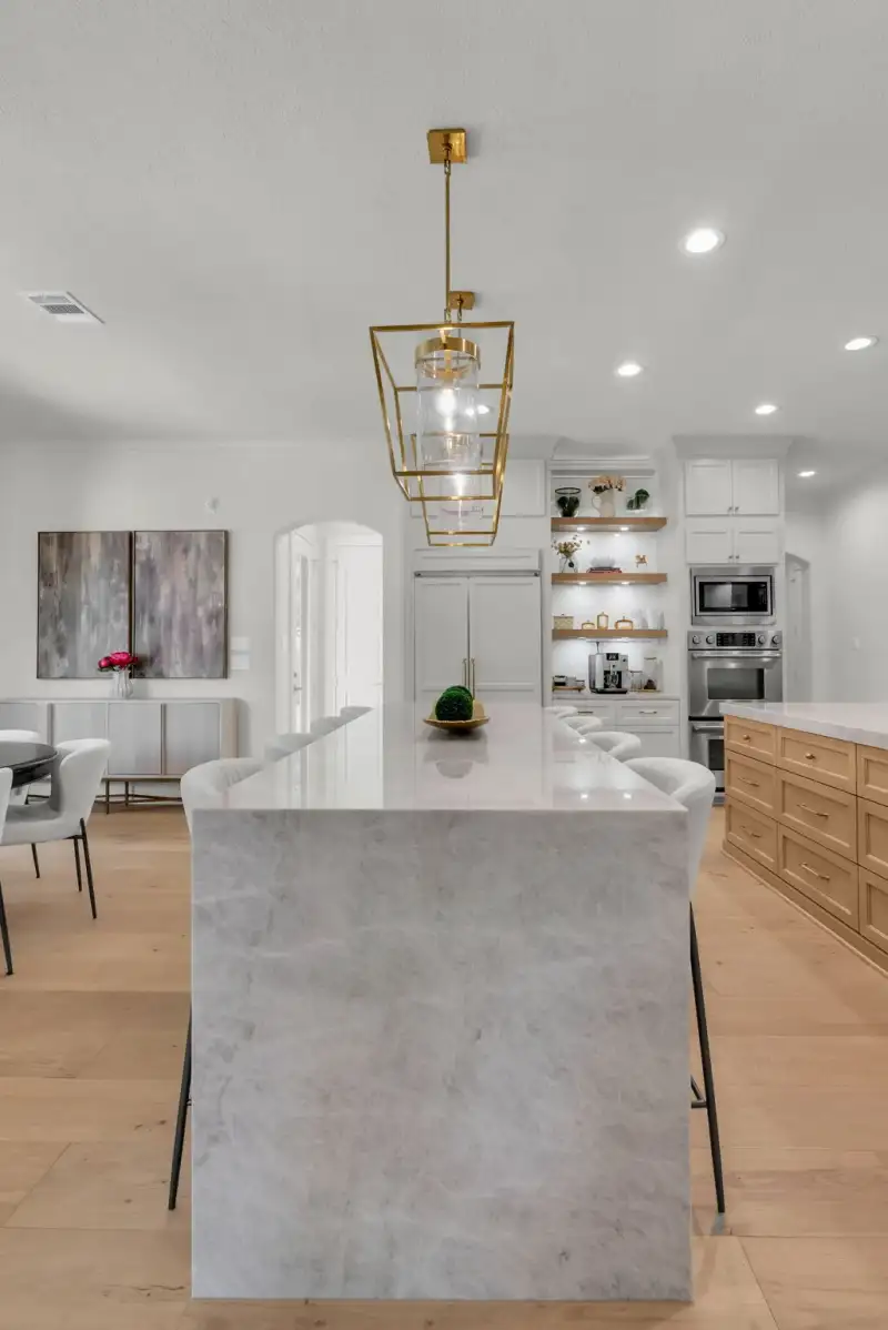 Kitchen island with a white marble-look waterfall countertop, bar stools, and stainless wall ovens and a microwave.