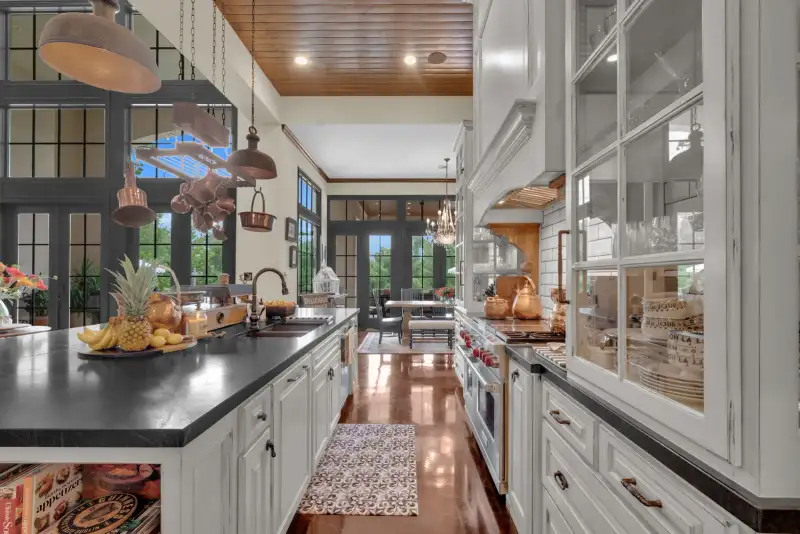 Kitchen with a dark countertop island, hanging copper pots, a wood-planked ceiling, and glass-front white cabinets.