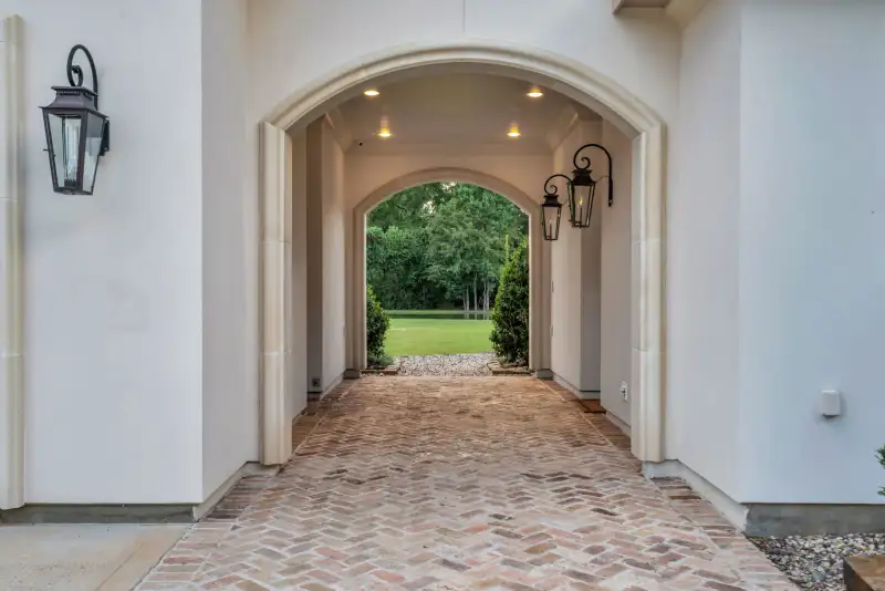 Arched brick passage with recessed ceiling lights and wall lanterns, opening onto a lawn.