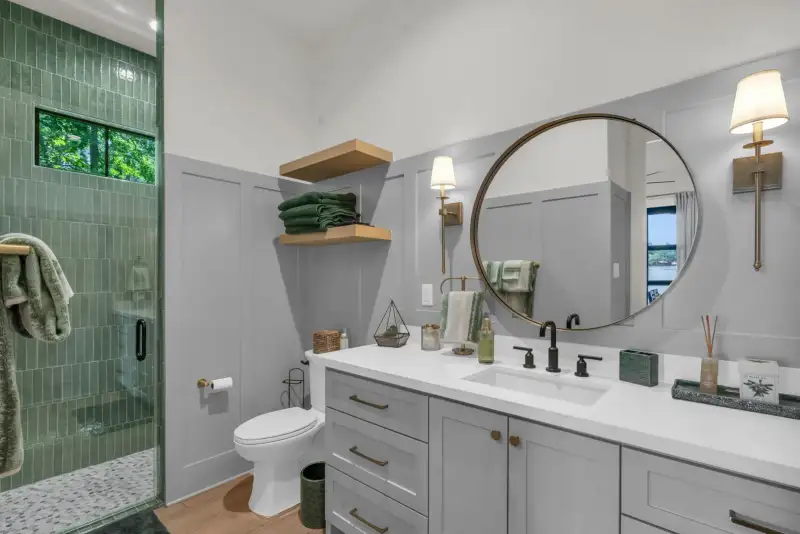 Bathroom with a green tiled shower, a round mirror, wall sconces, a gray vanity, and wood floating shelves.
