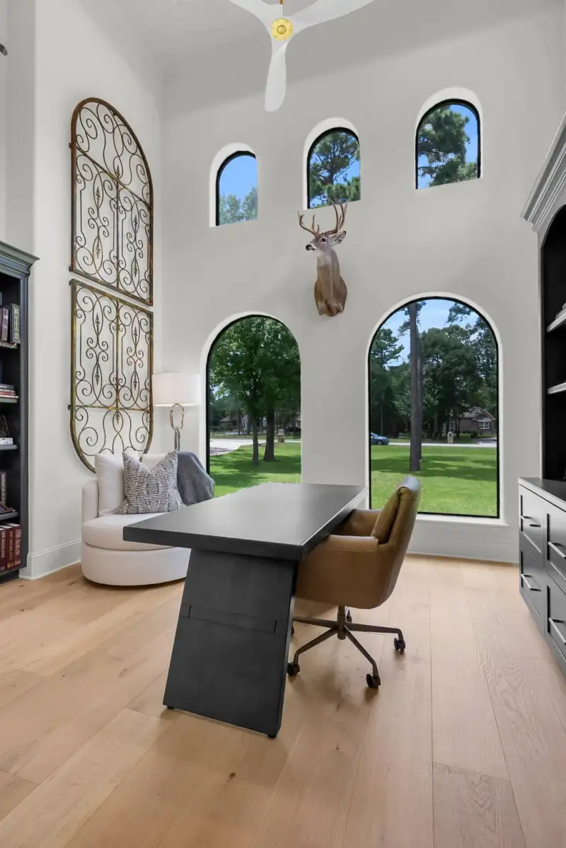 Study with a two-story ceiling, arched windows, a mounted deer head, and a dark desk.