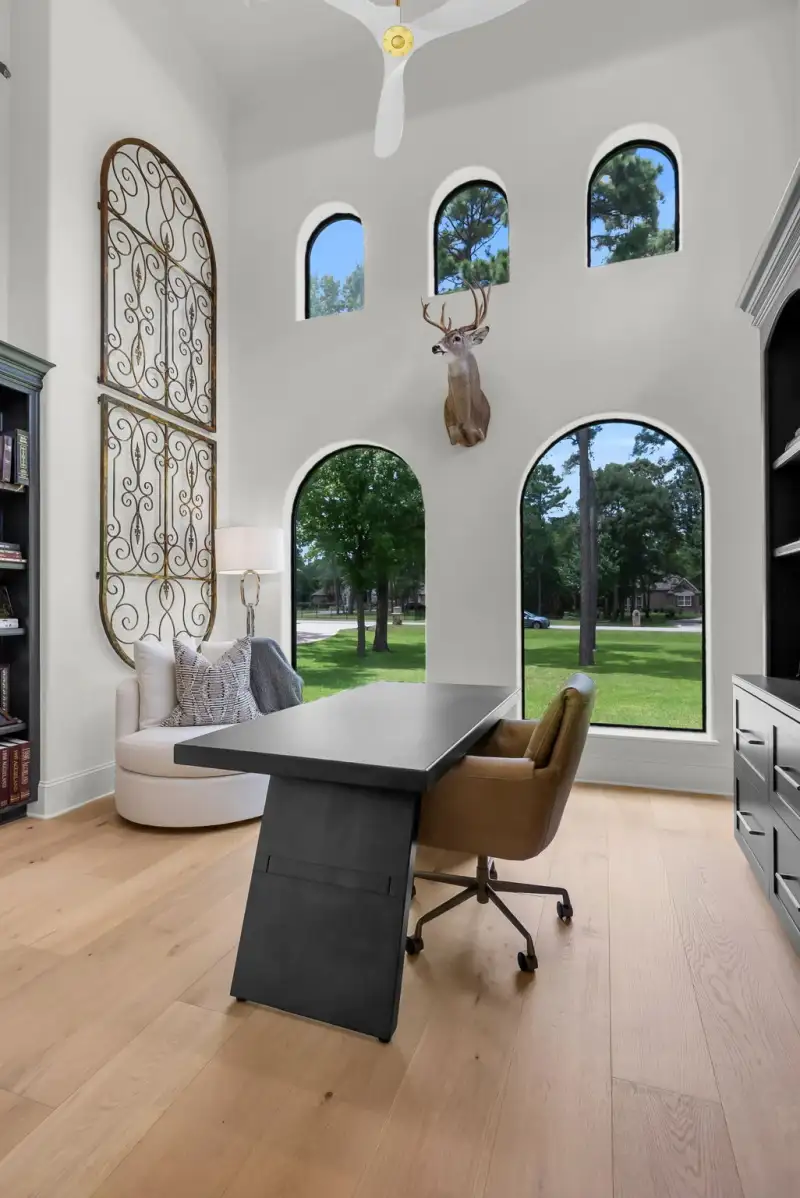 Study with a two-story ceiling, arched windows, a mounted deer head, and a dark desk.