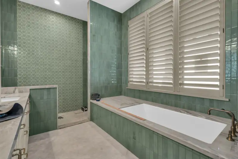 Shower with scalloped green tile, a tub, and a window with plantation shutters.