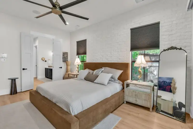 Bedroom with a brown upholstered bed, dark roller shades, and a tall arched mirror beside a window.