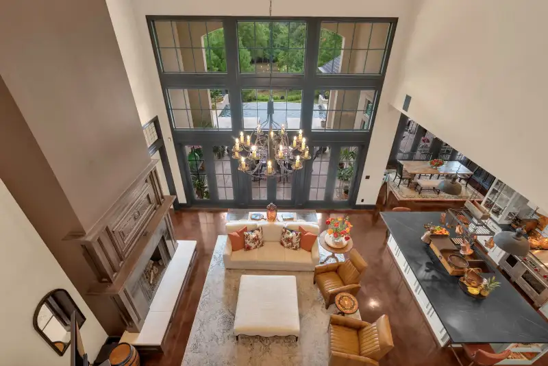 Two-story great room with a tall window wall, a cream ottoman, leather armchairs, and a dark island.