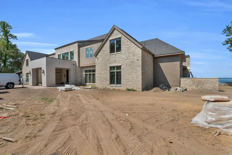 Ground-level view of the Escondido home's side wall with stone, tan lap siding, and a graded dirt yard.