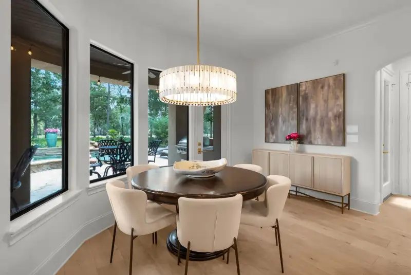 Dining area with a round dark table, upholstered chairs, a wood sideboard, and windows to the patio.
