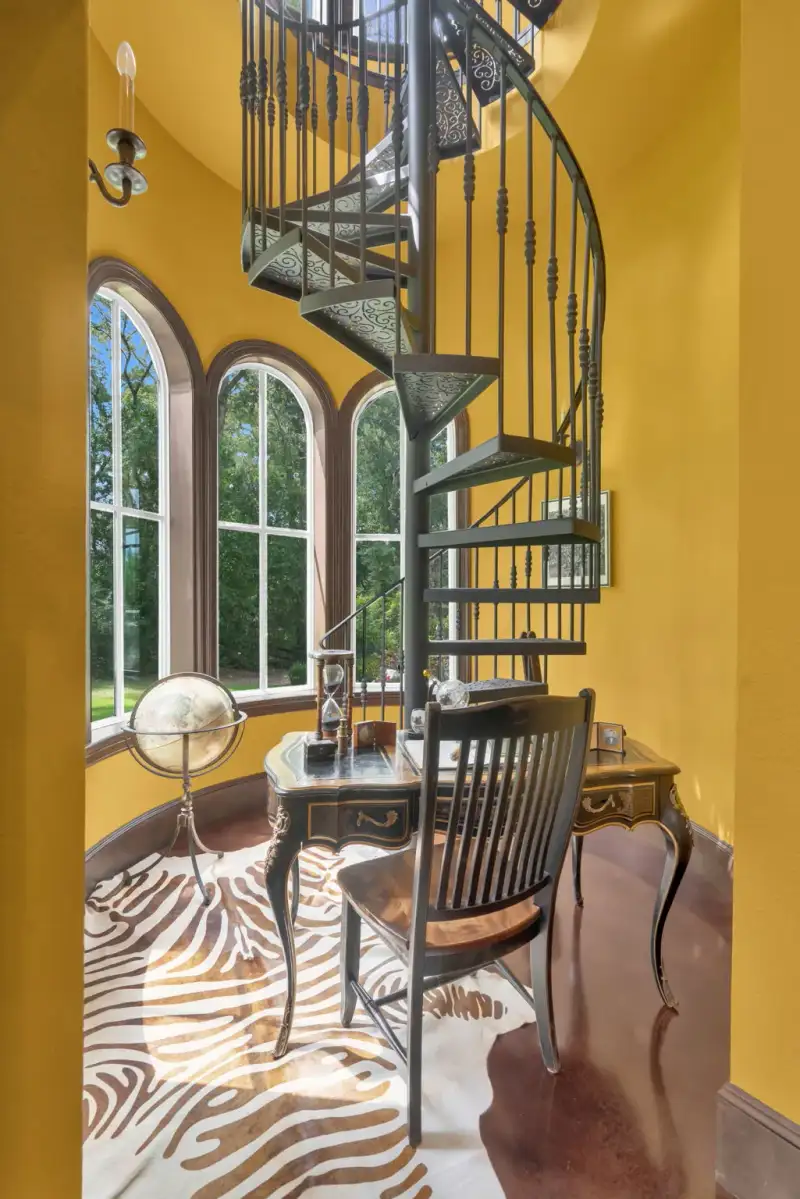 Iron spiral staircase in a yellow-walled turret beside arched windows, with an animal-hide rug and a wood writing desk.