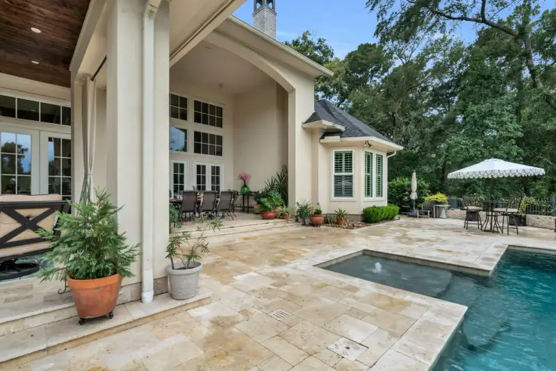 Pool deck beside a covered patio with white curtains, a dining set, potted plants, and a patio umbrella beyond.