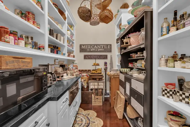 Pantry with white open shelving, canned goods, glass jars, hanging baskets, and a microwave on a dark counter.